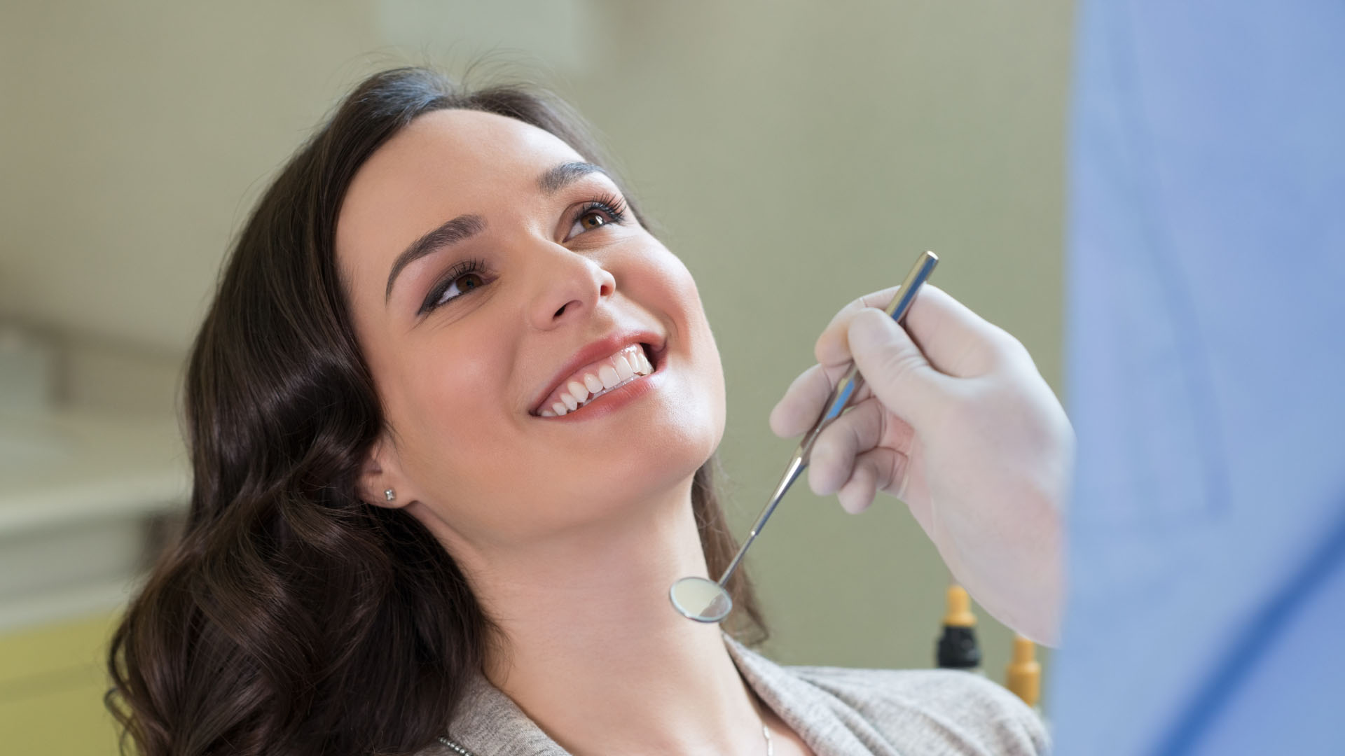 Smiling woman with healthy white teeth at a dental check-up, with a gloved dentist holding a mouth mirror nearby